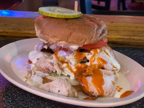 A stacked sandwich with grilled chicken, lettuce, tomato, and sauce, topped with a pickle slice on a toasted bun.