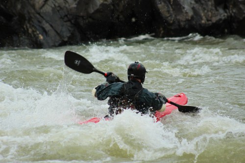 A person in a black helmet paddles a red kayak through turbulent, white-water rapids.