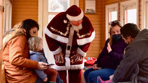 Santa Claus interacts with a family inside a cozy cabin, while a child sits on a woman's lap and others chat nearby.