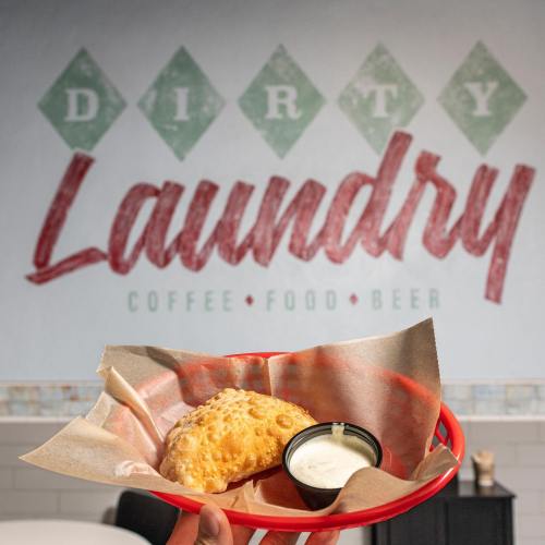 A hand holding a red plate with a golden pastry and a small bowl of dipping sauce, against a "Dirty Laundry" wall sign.