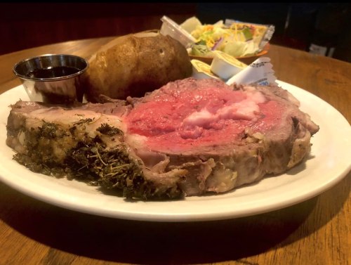 A plate featuring a juicy prime rib with herbs, a baked potato, and a side of dipping sauce.