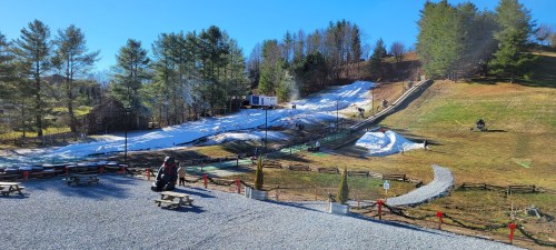 A snowy hill with people enjoying winter activities, surrounded by trees and a clear blue sky. Benches in the foreground.