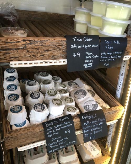 A display of various cheeses in a wooden shelf, with chalkboard labels showing prices and descriptions.