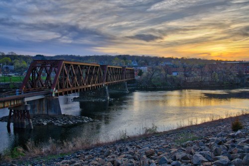 A rusty train bridge spans a calm river at sunset, with trees and buildings in the background under a cloudy sky.