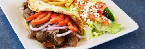 A plate featuring a pita filled with sliced meat, tomatoes, onions, fries, and a fresh salad with feta cheese.