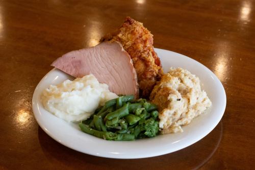 A plate with sliced ham, fried chicken, mashed potatoes, green beans, and stuffing on a wooden table.