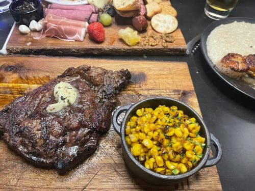 A juicy steak topped with butter, served with a side of sautéed corn and a charcuterie board in the background.