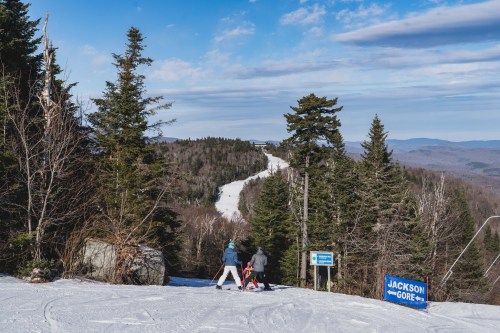 Two skiers walk towards a snowy slope surrounded by trees, with a sign for Jackson Gore in the foreground.