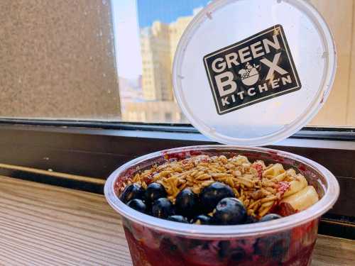 A clear container of acai bowl topped with granola and blueberries, with a logo for Green Box Kitchen in the background.