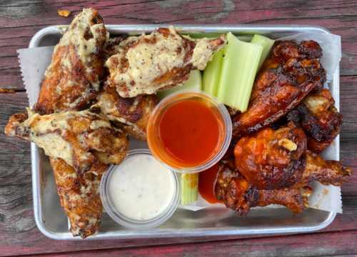 A metal tray with assorted chicken wings, celery sticks, and two dipping sauces: one red and one white.