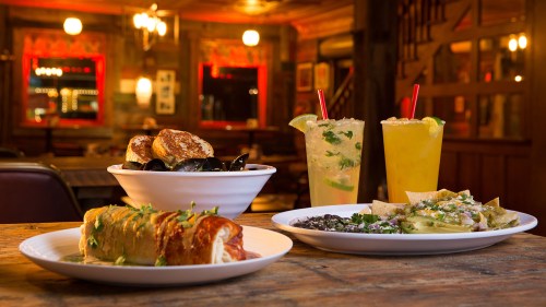A table with a variety of dishes: mussels, a burrito, nachos, and two colorful cocktails in a cozy restaurant setting.