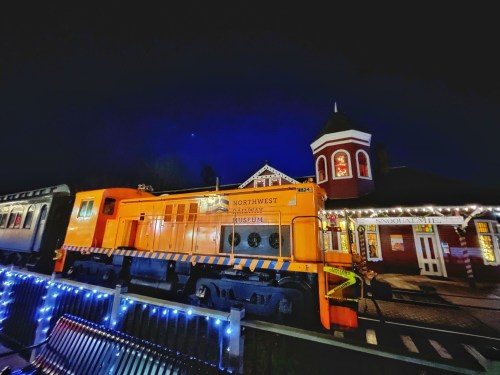 An orange train at night, illuminated by festive lights, next to a historic building with a tower.