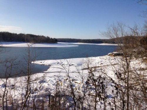 A serene winter landscape featuring a snow-covered shoreline and calm blue water under a clear sky.