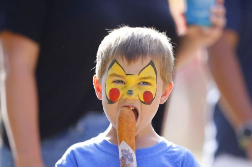 A boy with Pikachu face paint holds a corn dog, looking playfully at the camera.