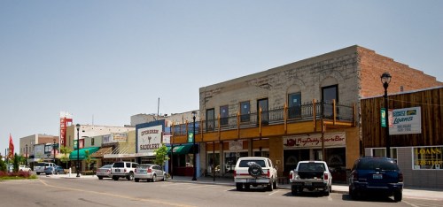 A small town street with shops, parked cars, and a clear blue sky. Buildings feature various signs and awnings.