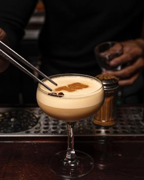 A bartender uses tweezers to place a garnish on a frothy cocktail in a coupe glass, set on a bar counter.
