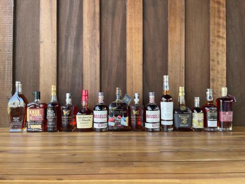 A collection of various whiskey bottles displayed on a wooden surface against a rustic wooden backdrop.