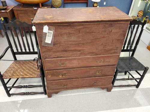 A vintage wooden chest of drawers flanked by two chairs, one with a woven seat and the other painted black.