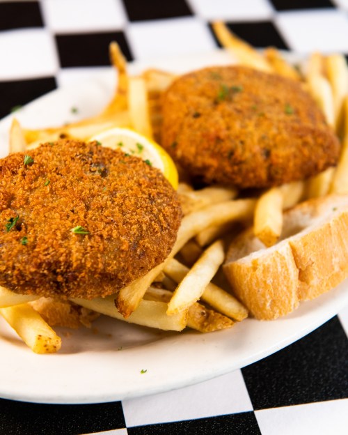Two golden-brown croquettes served with French fries and a slice of bread on a checkered tablecloth.