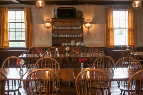 Cozy dining room with wooden tables, chairs, and floral centerpieces, illuminated by warm light from windows.