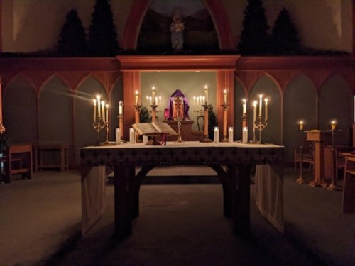 A dimly lit altar with candles, an open book, and a religious statue in the background, creating a serene atmosphere.