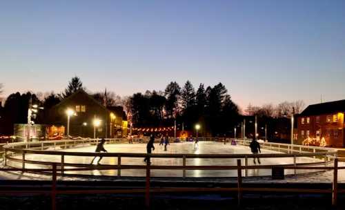 A winter scene of an outdoor ice skating rink at dusk, with skaters and festive lights around the area.