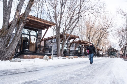 A person walks along a snowy path lined with modern cabins and bare trees in a winter landscape.