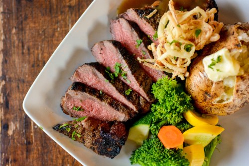 A plate of sliced grilled steak with a baked potato, sautéed vegetables, and crispy onion rings.