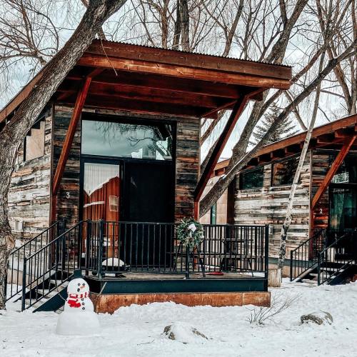 A cozy cabin with wooden exterior, surrounded by snow and trees, featuring a small snowman on the porch.