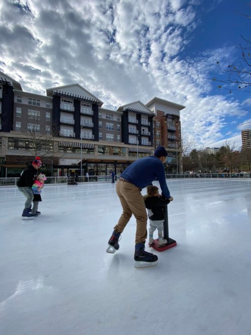 A man helps a child skate on an ice rink, while another skater glides in the background under a cloudy sky.