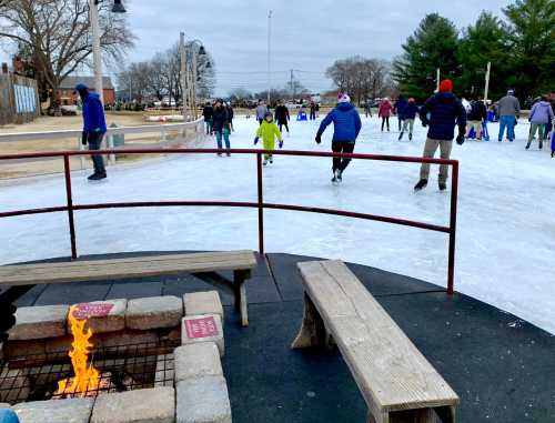 A winter scene of people ice skating on a rink, with a fire pit and benches in the foreground.