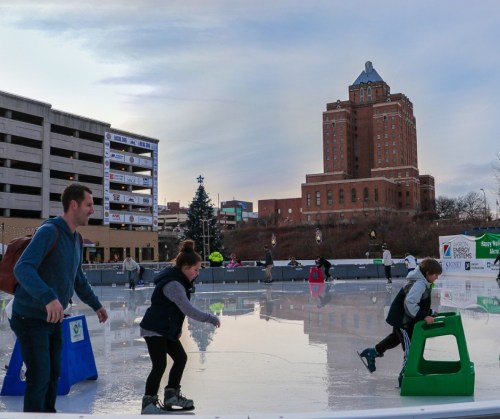 A family enjoys ice skating on an outdoor rink with a city skyline in the background.