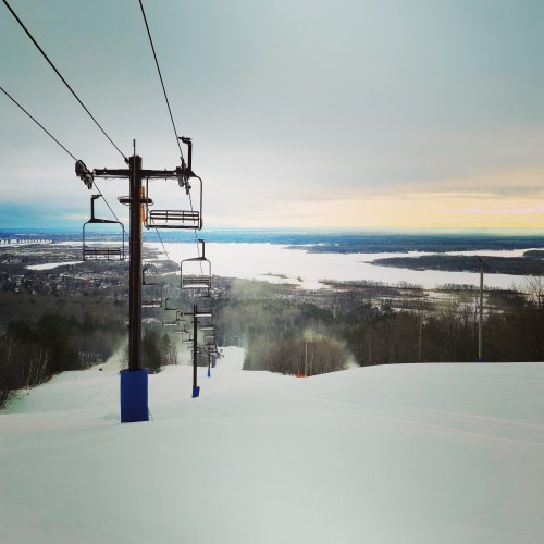 Ski lift towers over a snowy landscape, with a view of a lake and distant hills under a cloudy sky.