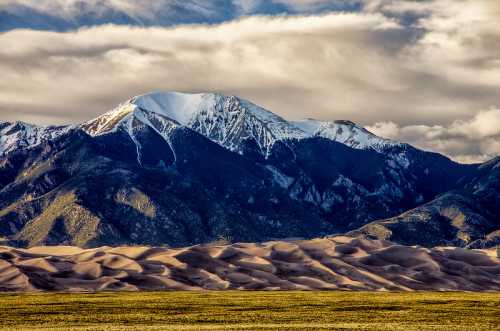 Snow-capped mountains rise behind rolling sand dunes under a cloudy sky, showcasing a striking natural landscape.