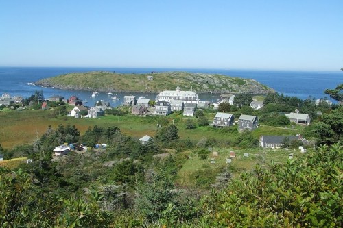 A scenic view of a coastal village with houses, greenery, and an island in the background under a clear blue sky.