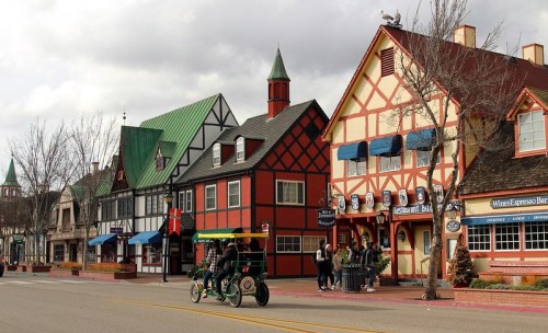 Colorful, European-style buildings line a street, with a bike taxi passing by and trees in the foreground.