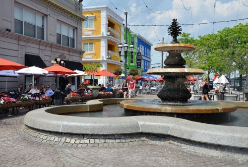 A vibrant plaza featuring a decorative fountain, outdoor seating, colorful buildings, and umbrellas on a sunny day.