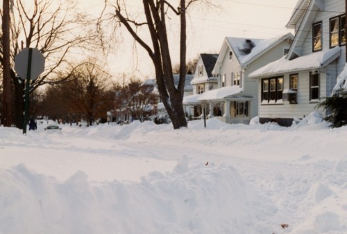 A snowy residential street lined with houses, trees, and a stop sign, creating a serene winter scene.