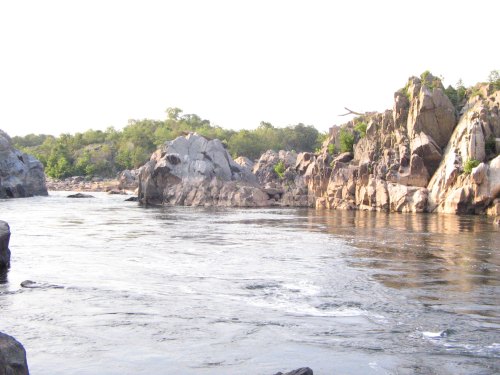 A serene river flows between rocky cliffs, surrounded by lush greenery under a bright sky.