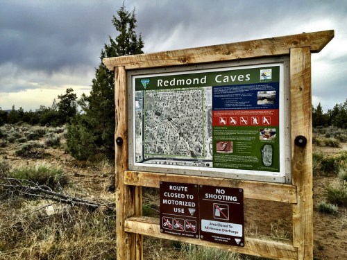 Sign for Redmond Caves with a map and information, surrounded by a natural landscape under a cloudy sky.