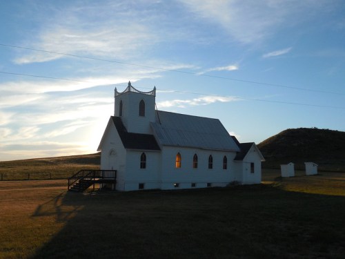 A white church with a steeple stands against a sunset, surrounded by open fields and a distant hill.