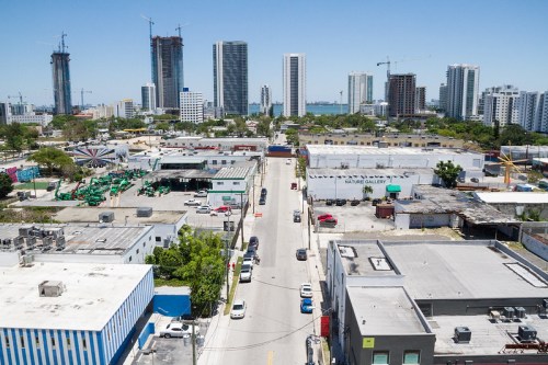 Aerial view of a cityscape with tall buildings, construction sites, and a street lined with low-rise buildings and vehicles.