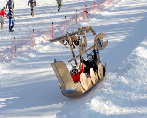 Two children in a cardboard boat with sails and oars, joyfully sliding down a snowy hill.
