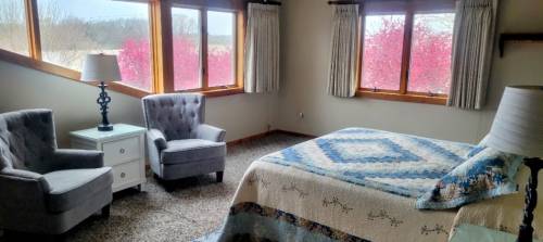 Cozy bedroom with a quilted bed, two armchairs, and large windows showcasing blooming pink trees outside.