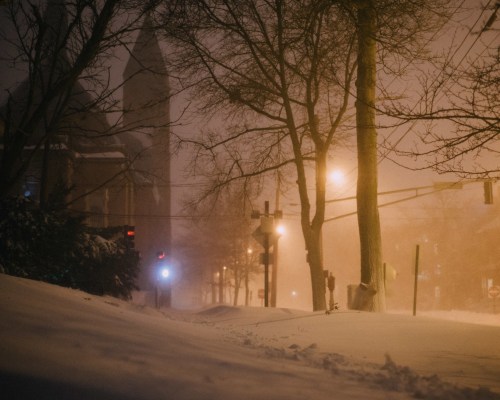 A snowy street scene at night, with trees, traffic lights, and a church in the background, shrouded in fog.
