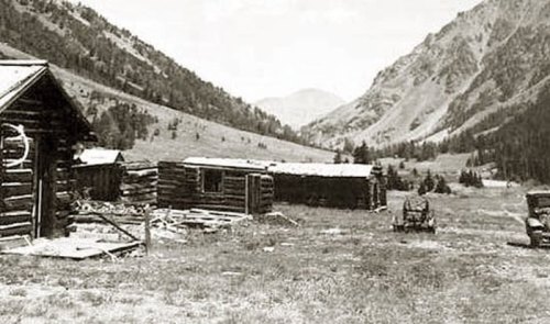 Black and white photo of rustic cabins in a mountainous landscape, with a vintage vehicle and open fields.