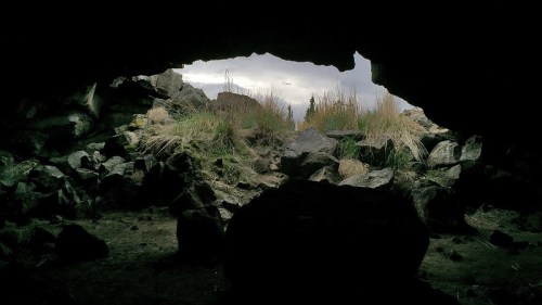 View from inside a dark cave, showing a rocky entrance with grass and a cloudy sky in the background.