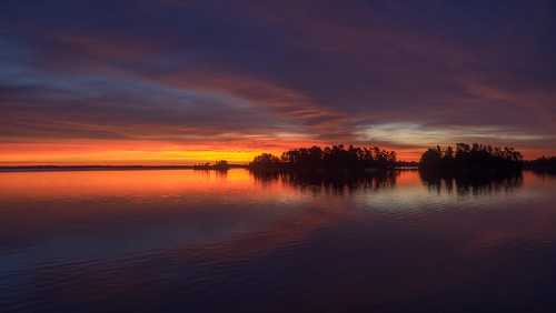 A serene lake at sunset, with vibrant orange and purple skies reflecting on the water and silhouetted trees on the horizon.