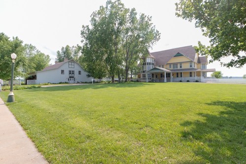 A spacious green lawn with a large yellow house and a white barn surrounded by trees under a clear sky.