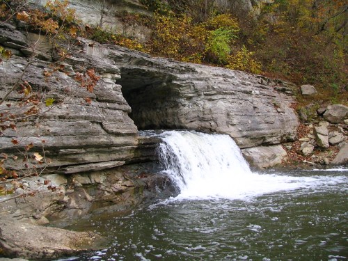 A small waterfall cascades over rocky terrain into a serene river, surrounded by autumn foliage.
